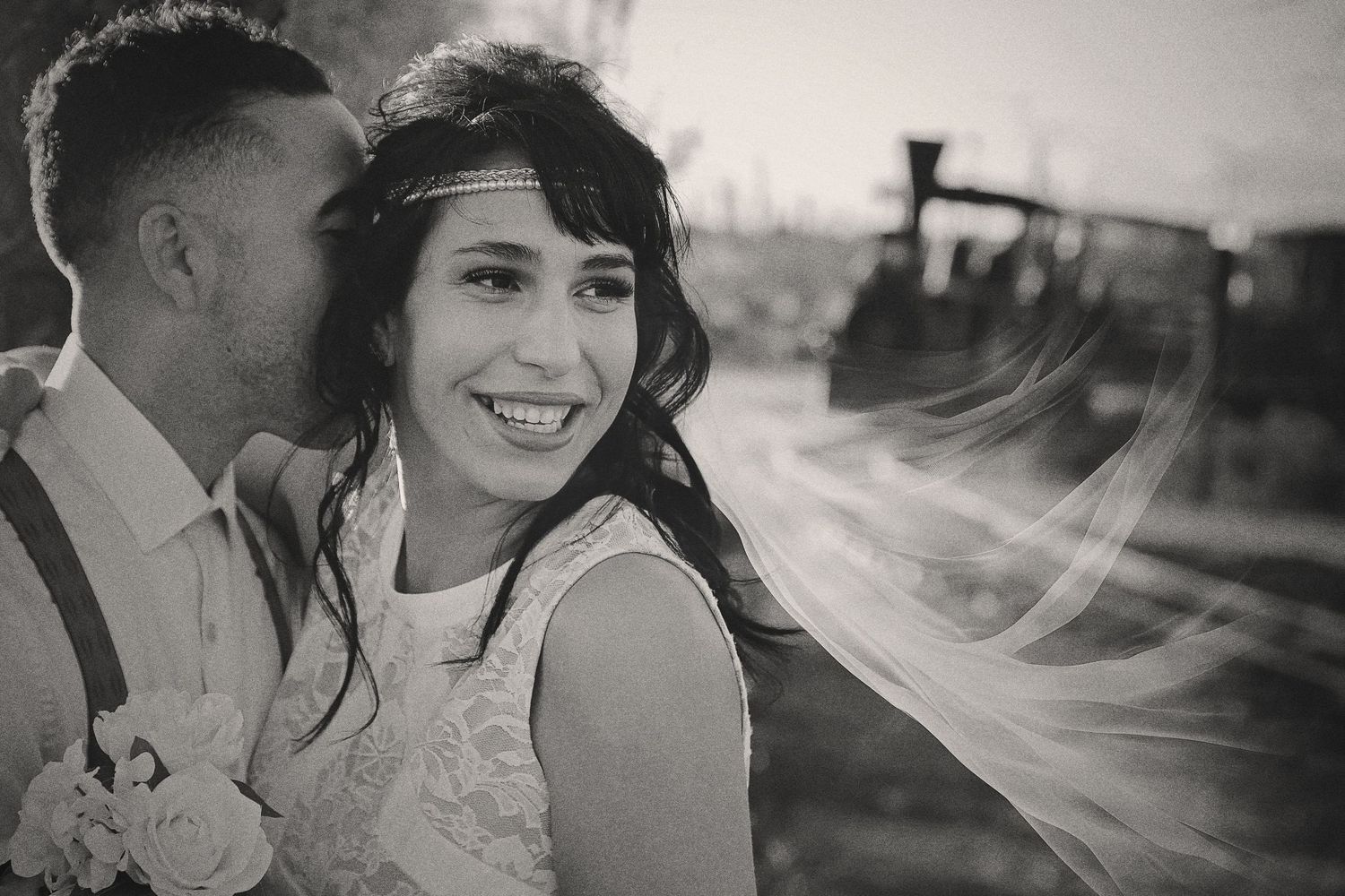 Black and white photo of a smiling bride with a flowing veil as her groom in suspenders kisses her cheek near an old train.