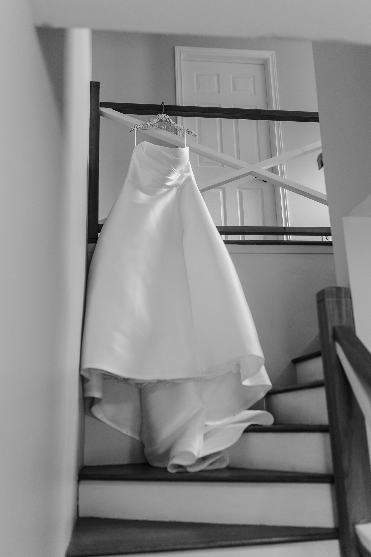 Strapless white wedding dress with a long train hanging from a railing above a wooden staircase in black and white.