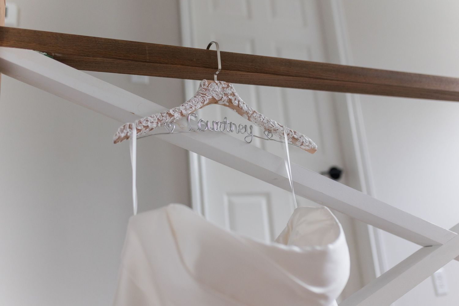 White wedding dress hanging on a decorative beaded hanger from a wooden beam in a bright room with white walls and a door.