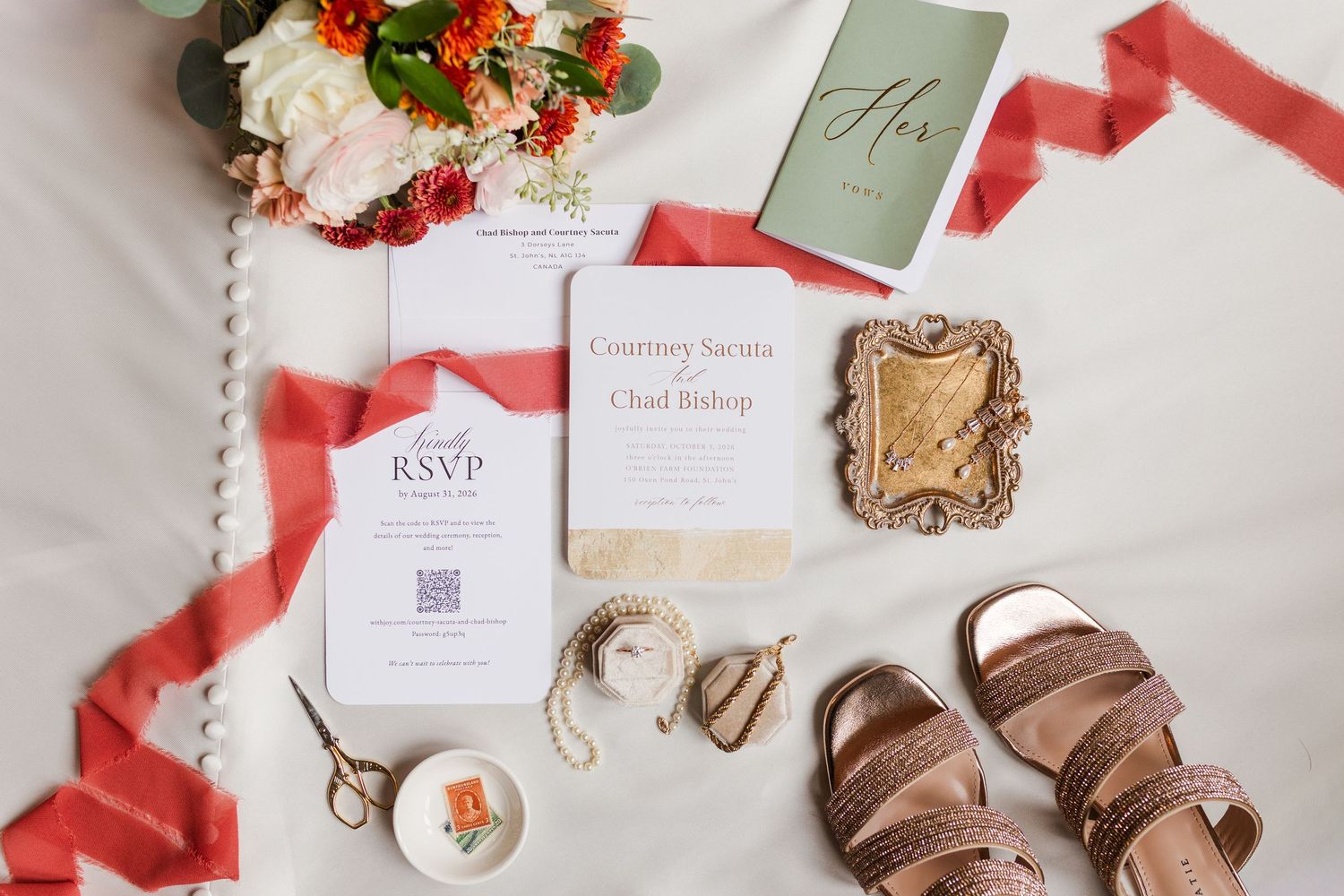 Flat lay of a wedding invitation suite with RSVP card, red ribbon, orange floral bouquet, blush sandals, scissors and a decorative frame.