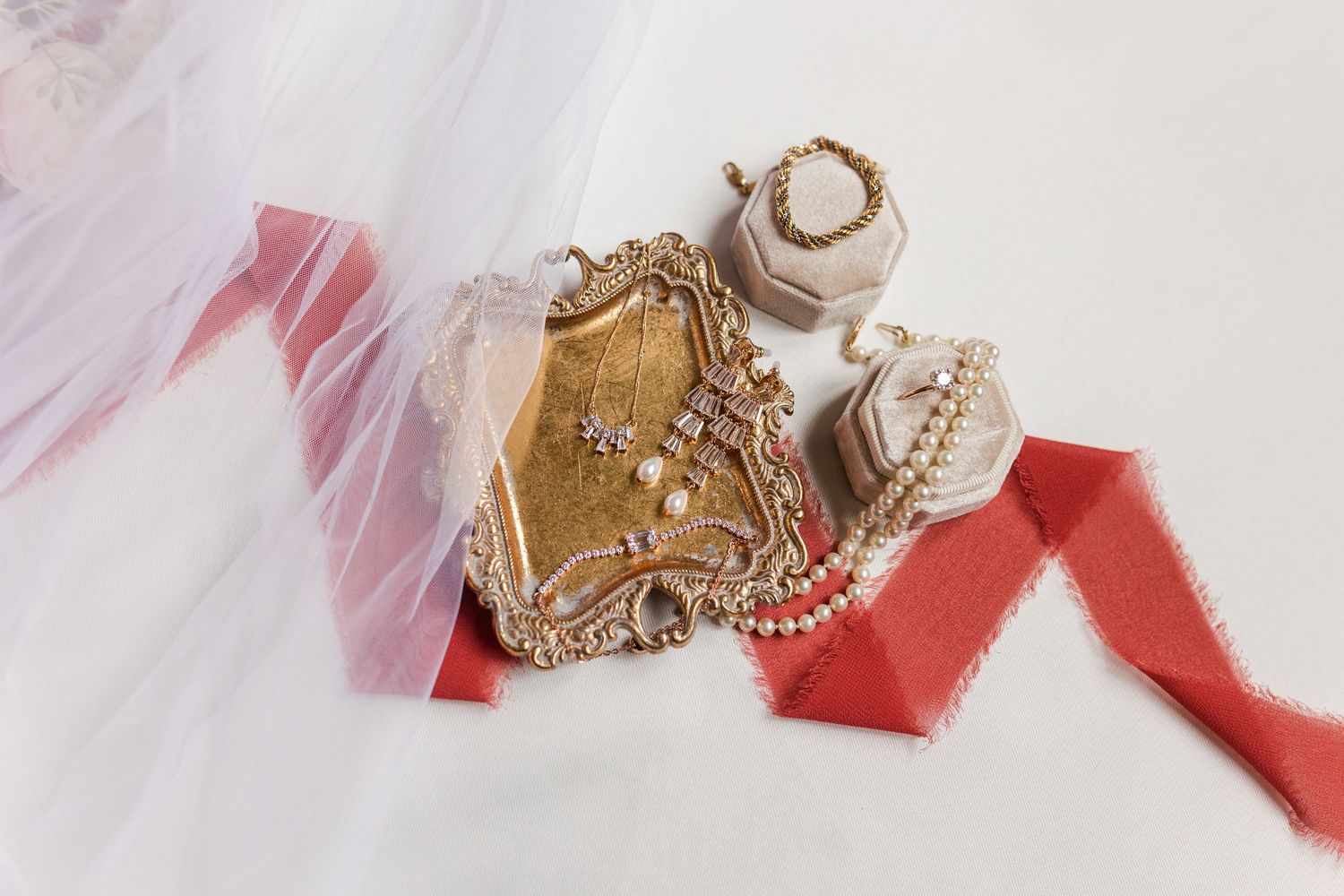Vintage gold ornate tray with pearl jewelry and rings in blush boxes on a red silk ribbon beside a sheer white veil.