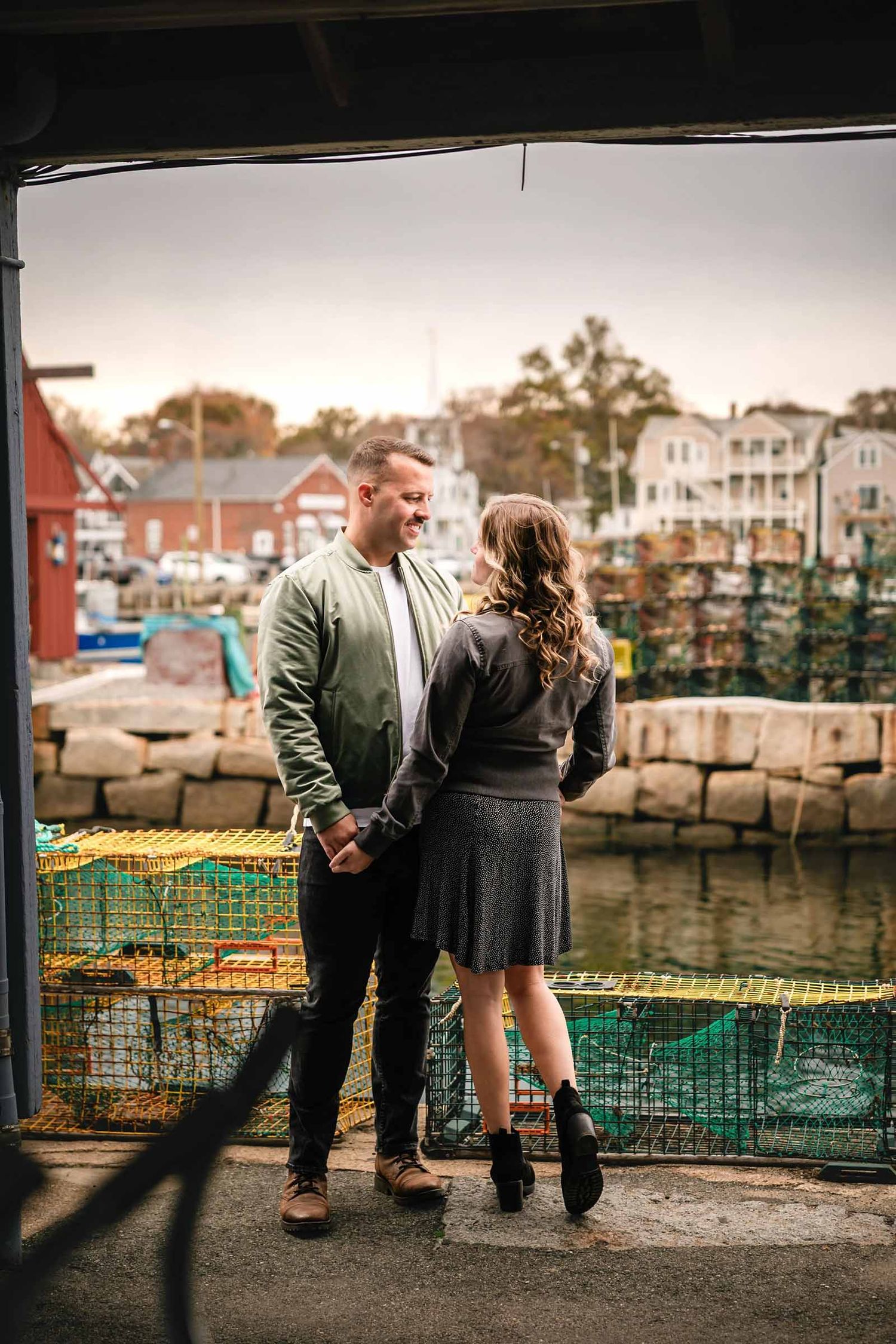 Couple standing together at a waterfront railing, gazing toward the horizon in quiet connection.  Engagement at Rockport Harbor.