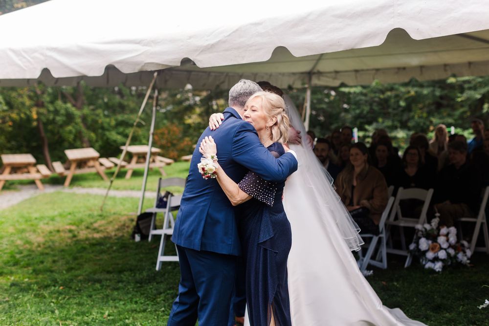 A bride in a white wedding gown embraces a man in a navy suit during an outdoor tent ceremony as guests watch from white chairs.