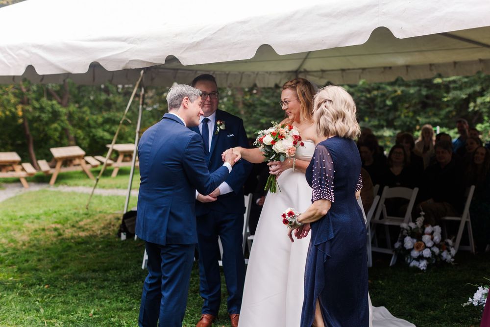 A father in a navy suit hands his bride in a white gown to the groom during an outdoor tent wedding ceremony as a woman and guests look on.
