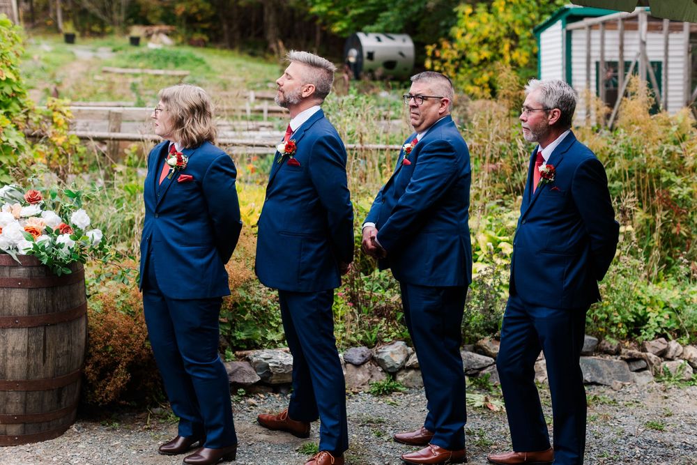 Four groomsmen in navy suits with red boutonnieres stand in a row at an outdoor fall wedding beside a wooden barrel of flowers.