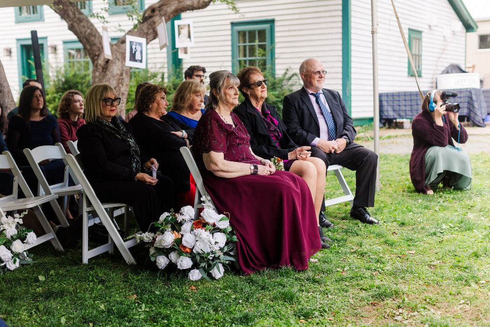Wedding guests seated on white folding chairs on a lawn watch an outdoor ceremony, with a white floral arrangement in the foreground.