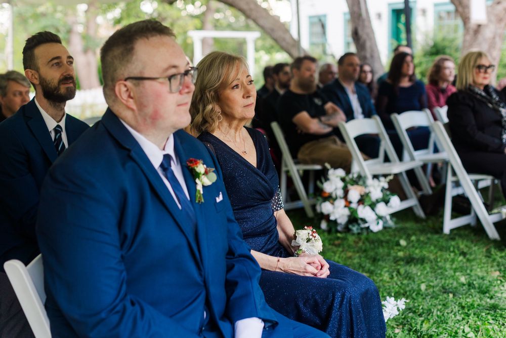 Man in navy suit with boutonniere and woman in blue dress sit among guests during an outdoor garden wedding ceremony with white flowers.