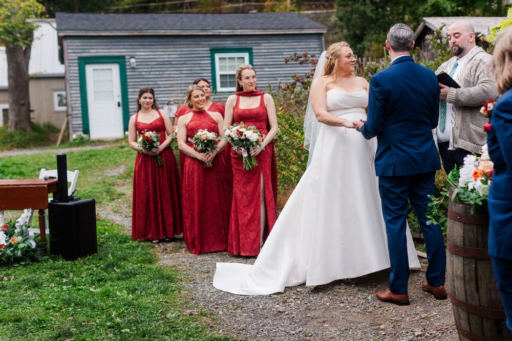 A bride in a white gown and a groom in a navy suit exchange vows outdoors as bridesmaids in red dresses hold bouquets nearby.