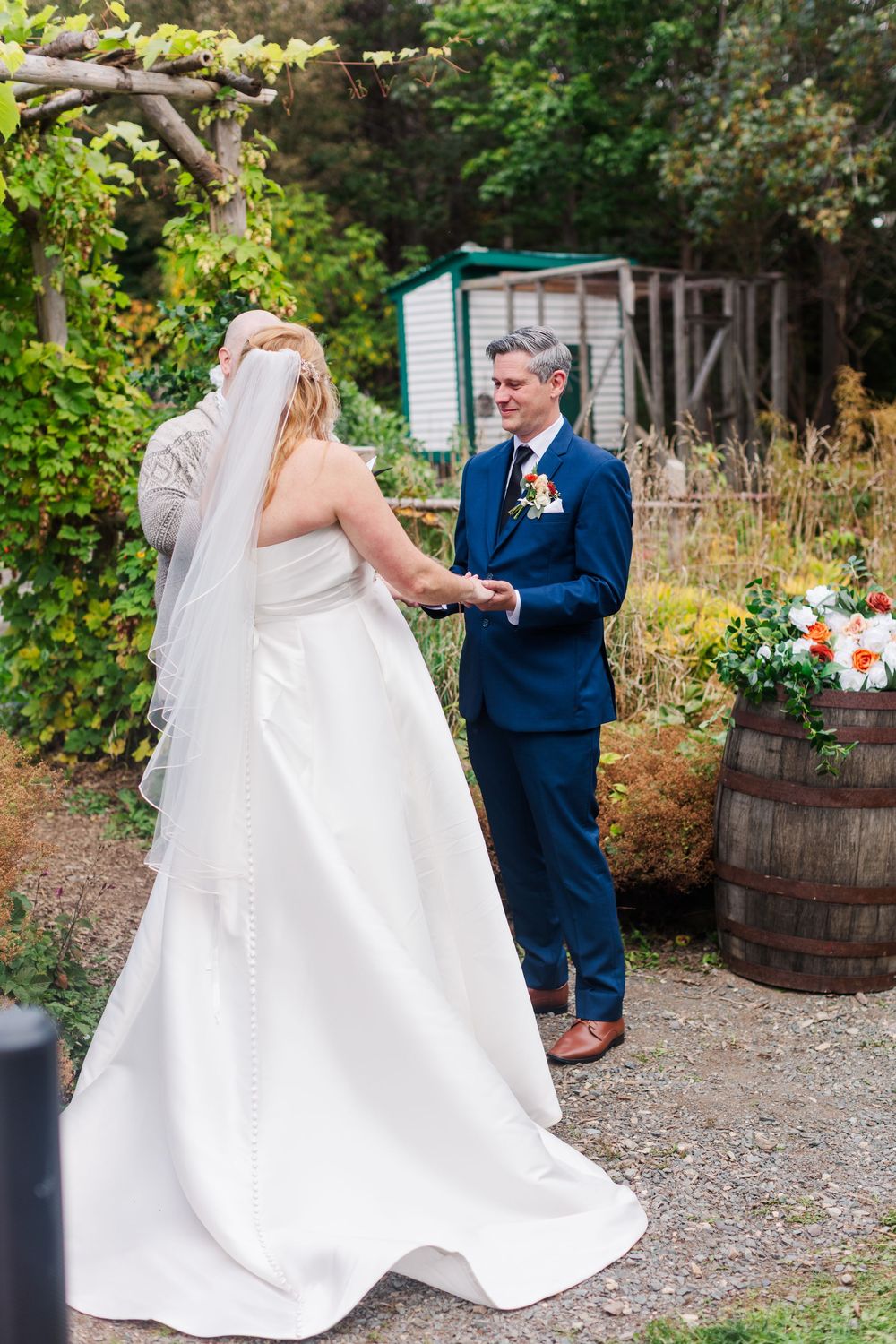 Bride in a white gown and veil holds hands with a groom in a navy suit during an outdoor garden wedding ceremony.