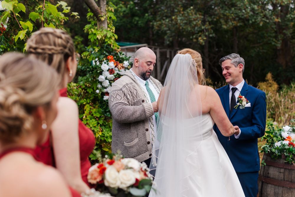 A smiling groom in a navy suit holds hands with his veiled bride as an officiant in a tweed jacket leads their outdoor wedding ceremony.