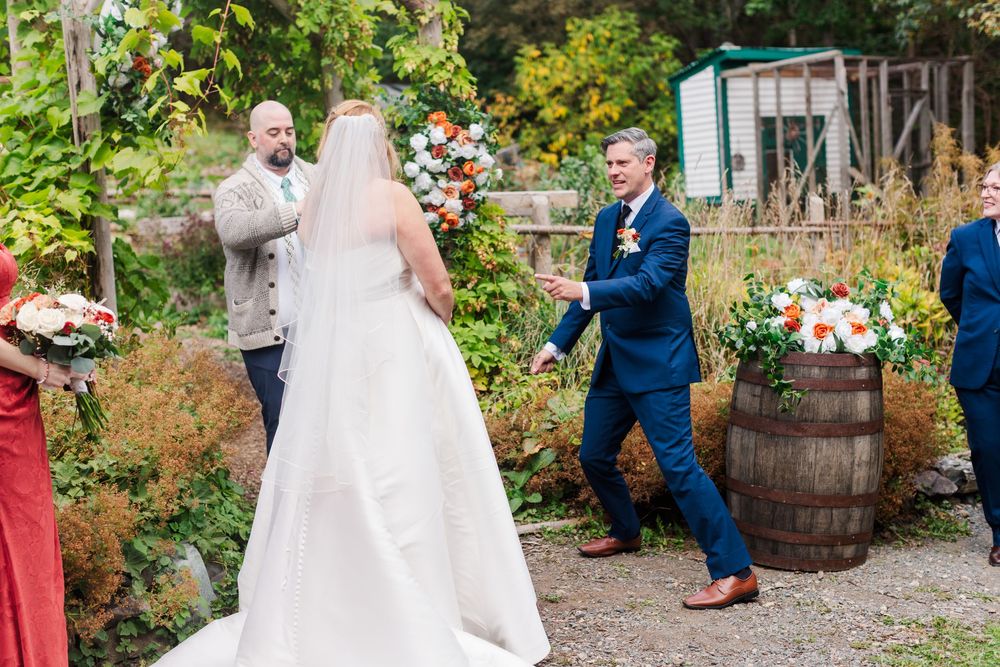 Bride in a white gown faces the groom and officiant during an outdoor garden wedding ceremony with floral arrangements and a wooden barrel.