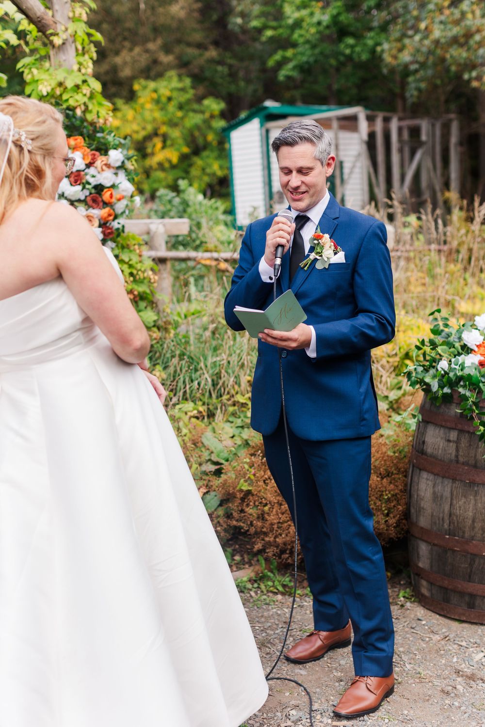 Groom in a navy suit reads vows from a green book into a microphone during an outdoor garden wedding with floral barrel decor.