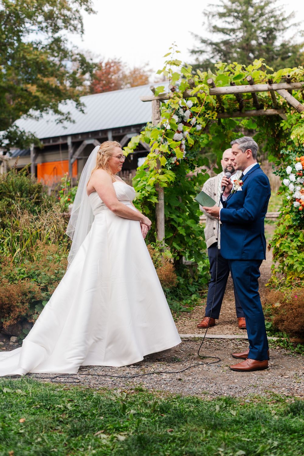 Bride in a white gown and veil faces a groom in a navy suit as an officiant speaks beneath a vine-covered arbor at an outdoor wedding.