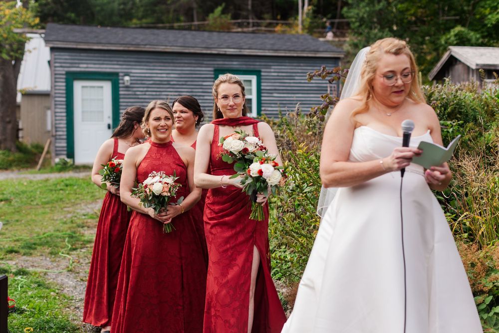 Bride in a white strapless gown speaks into a microphone while bridesmaids in red dresses hold bouquets at an outdoor wedding ceremony.