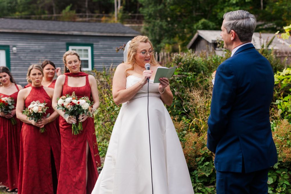 Bride in a white gown reads her vows from a card at an outdoor garden wedding as bridesmaids in red dresses hold bouquets nearby.