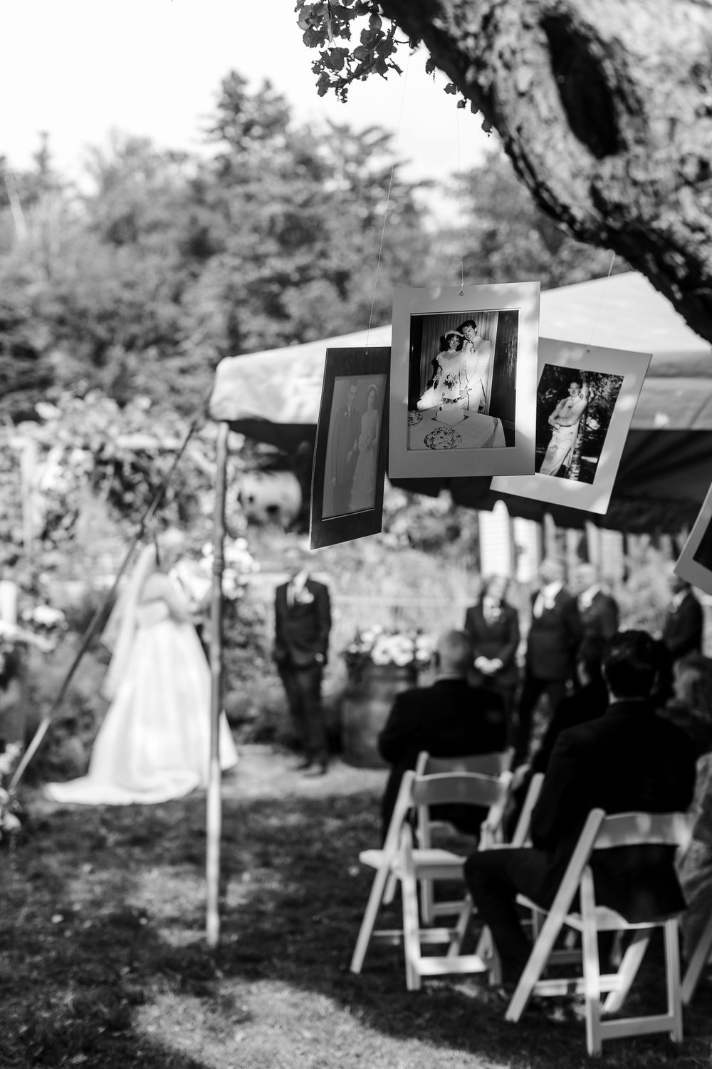 Black and white photo of an outdoor garden wedding ceremony with hanging framed family photos, a bride in a gown, and seated guests.