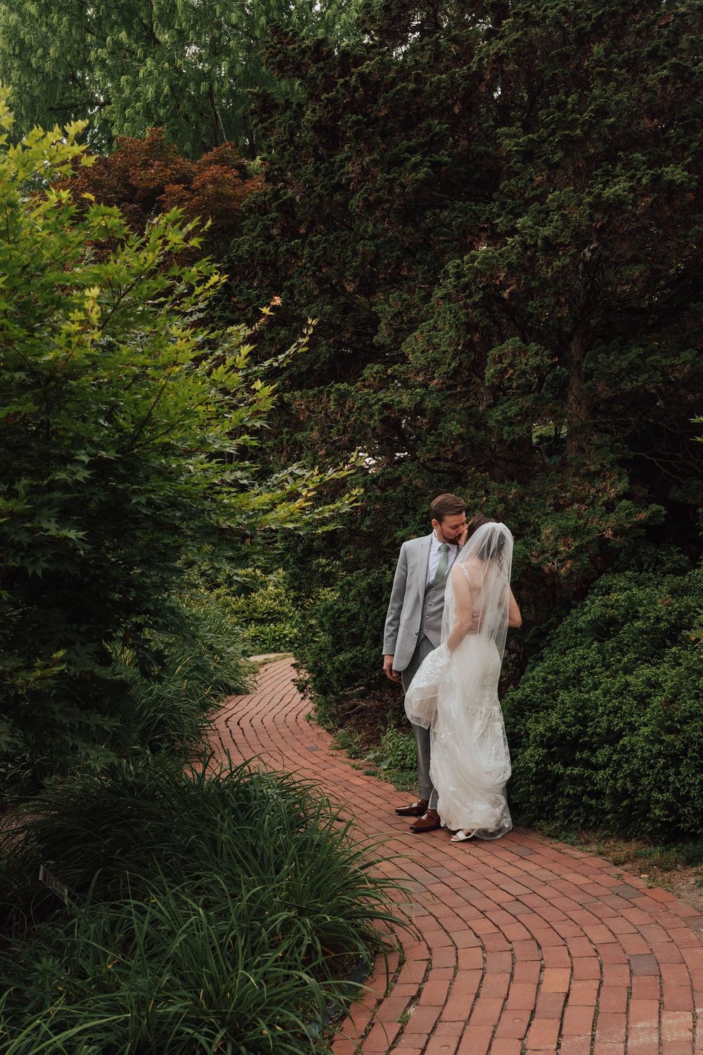 Bride and groom walking hand in hand on a brick garden path surrounded by lush green trees and shrubs.