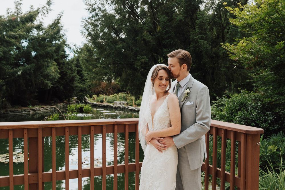 Bride and groom embracing on a wooden bridge surrounded by lush greenery and a serene pond at their outdoor wedding.