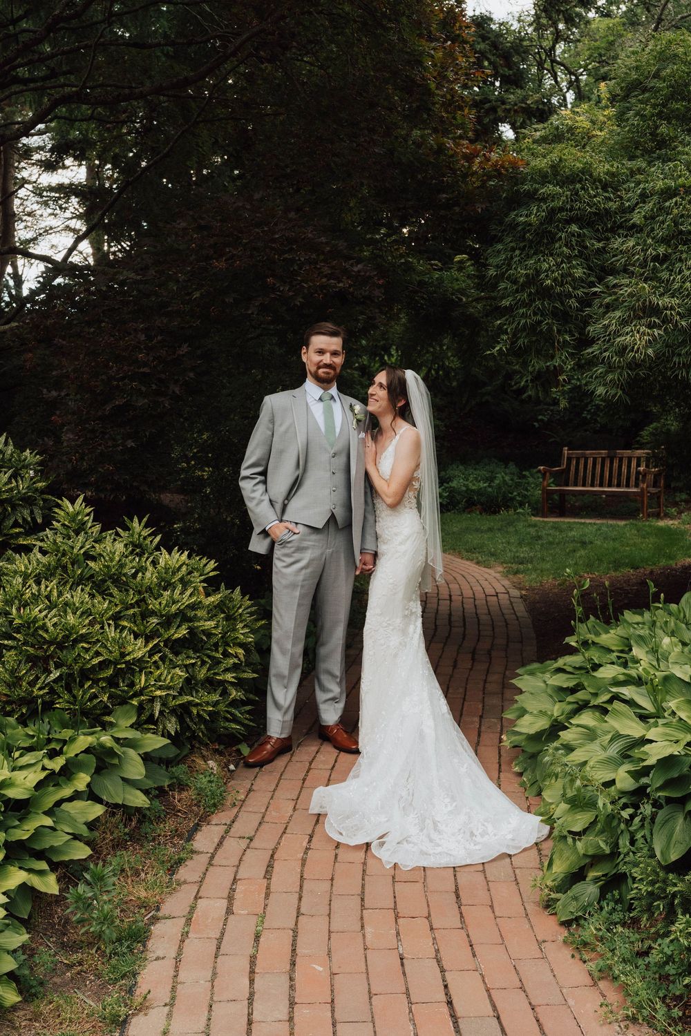 Bride in lace gown and groom in gray suit pose on brick garden path surrounded by lush greenery.