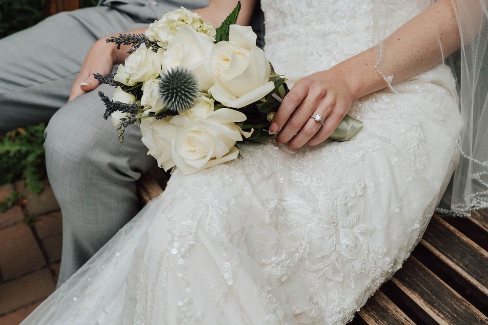 Bride in lace wedding dress holding white rose and thistle bouquet while seated beside groom in gray suit.