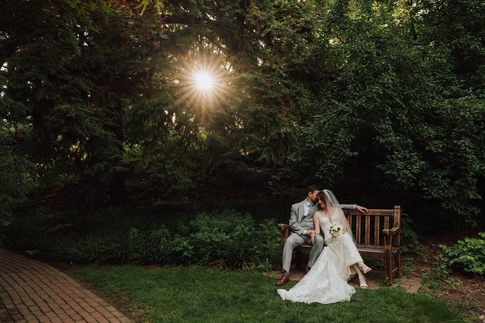 Bride and groom sit on a wooden bench in a lush garden, with sunlight bursting through the trees behind them.