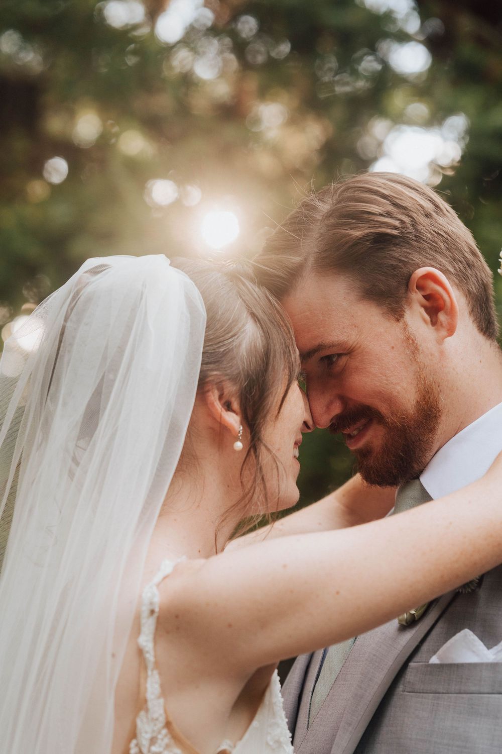 A bride and groom touch foreheads tenderly outdoors, bathed in golden sunlight, as she wears a flowing veil on their wedding day.