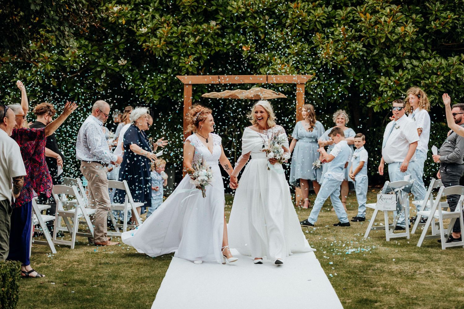 Bride and bride walking down the aisle at Hilton Lake Taupō, surrounded by confetti and smiling guests.