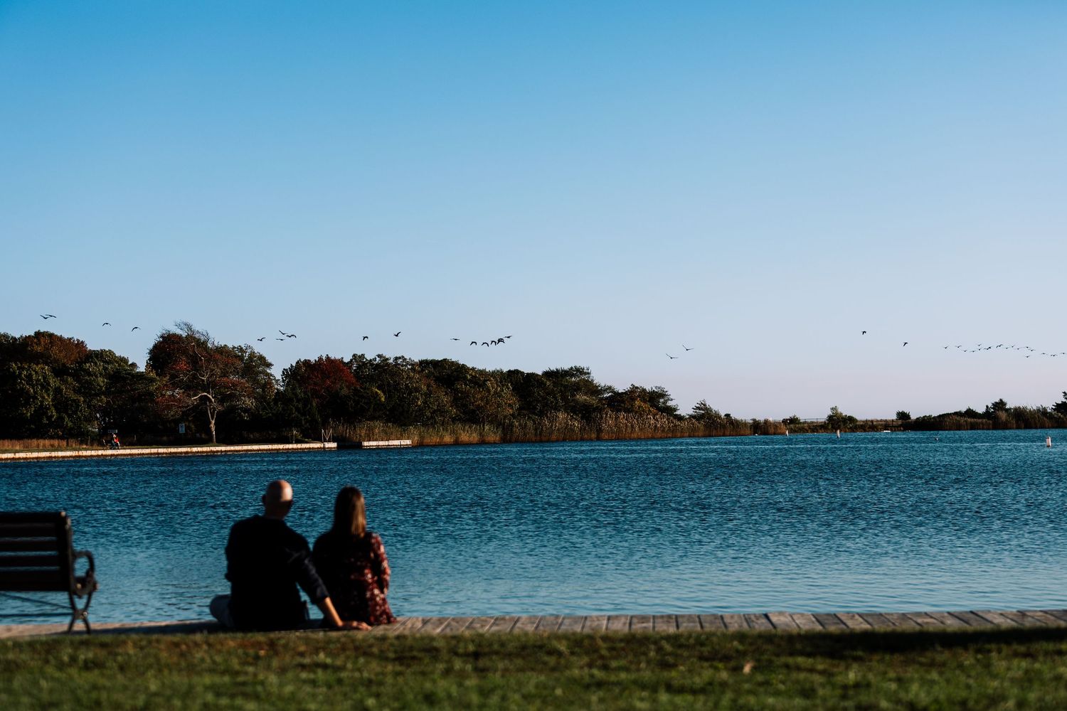 Couple sits together by the water at sunset watching a flock of birds fly over a calm bay lined with autumn trees.