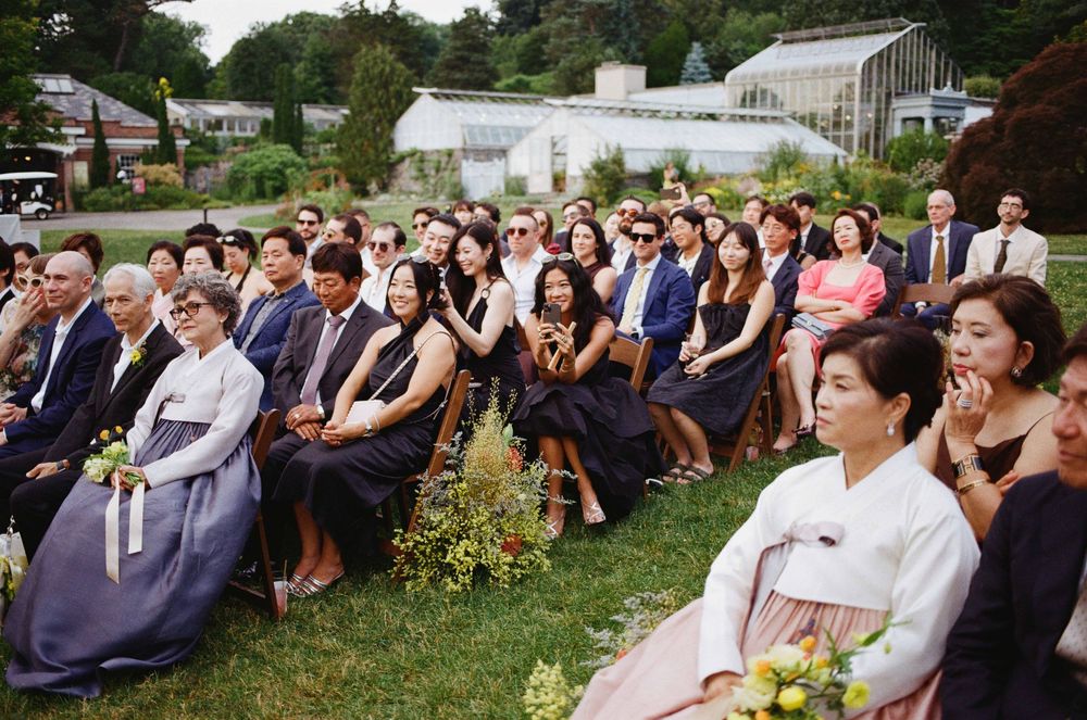 Guests seated outdoors at an elegant garden wedding ceremony, many wearing formal attire including traditional Korean hanbok.