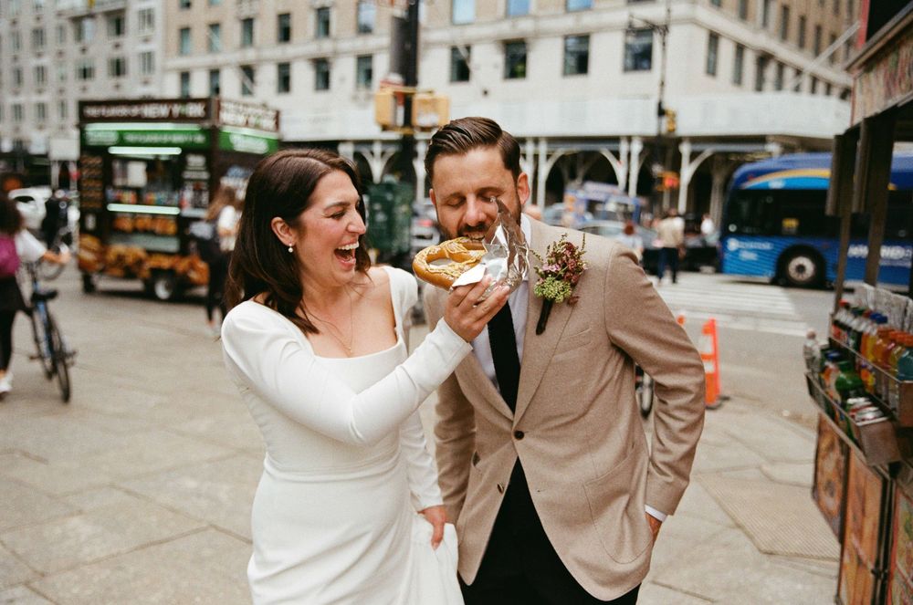 Bride and groom sharing a sandwich on a city street, laughing together in wedding attire outside urban buildings.