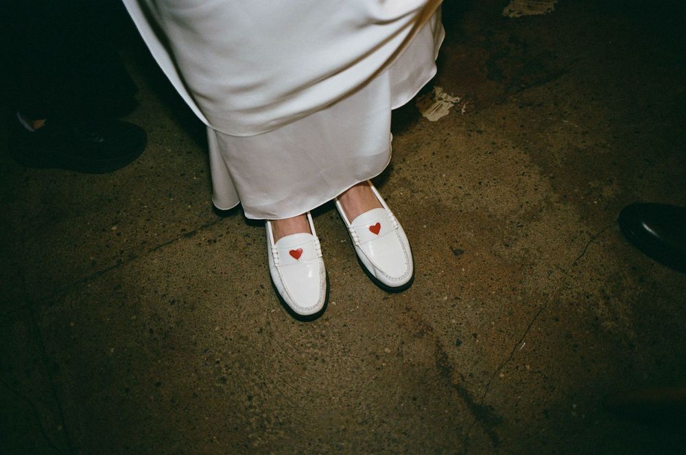 White loafers with red heart detail worn with a white satin dress on a stone floor.