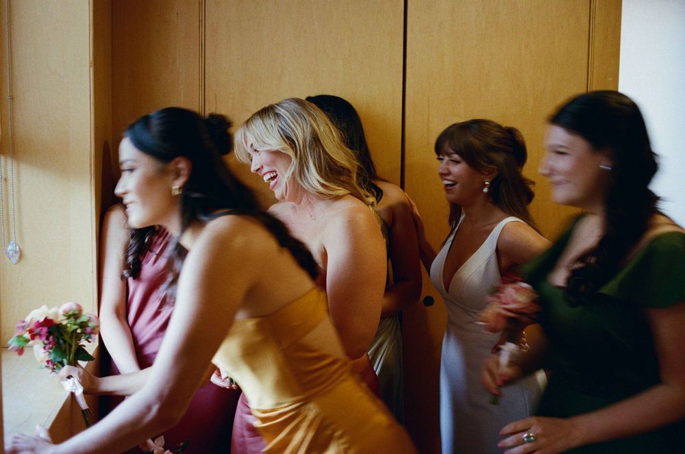 Bridesmaids in colorful satin dresses laughing together before a wedding ceremony indoors.