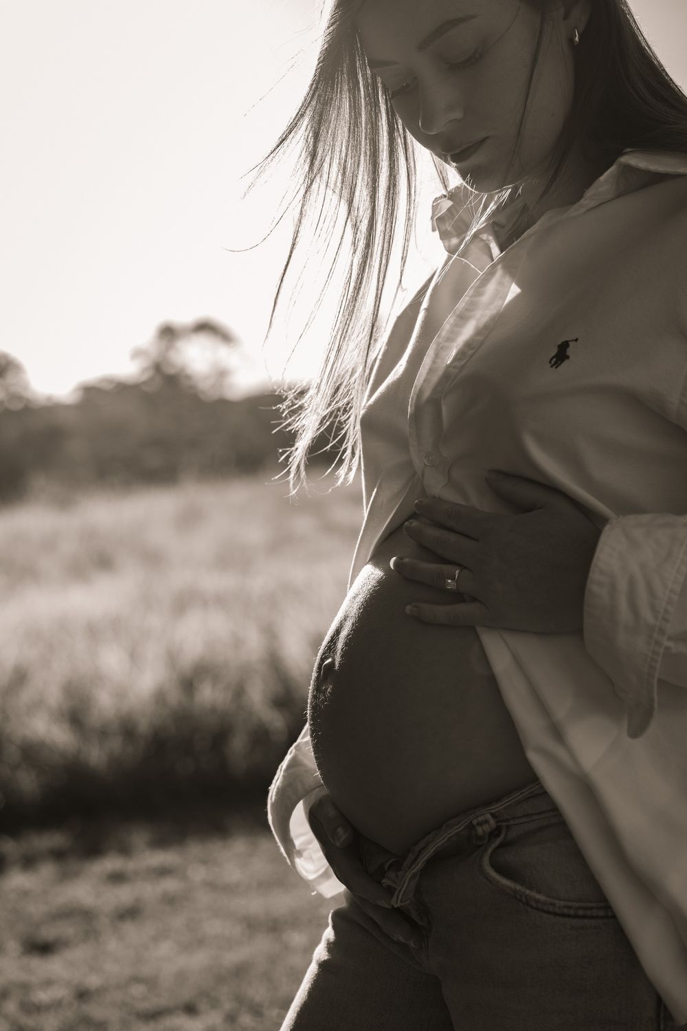 Black and white maternity portrait of a woman in a white shirt and jeans cradling her baby bump outdoors.