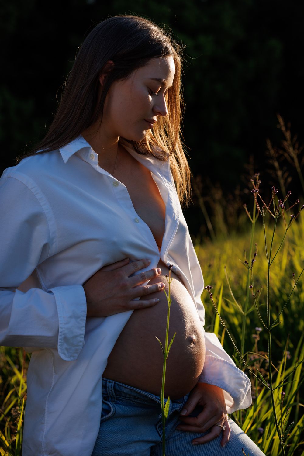 Pregnant woman in an open white shirt and jeans gently touches her belly in a sunlit grassy field at golden hour.