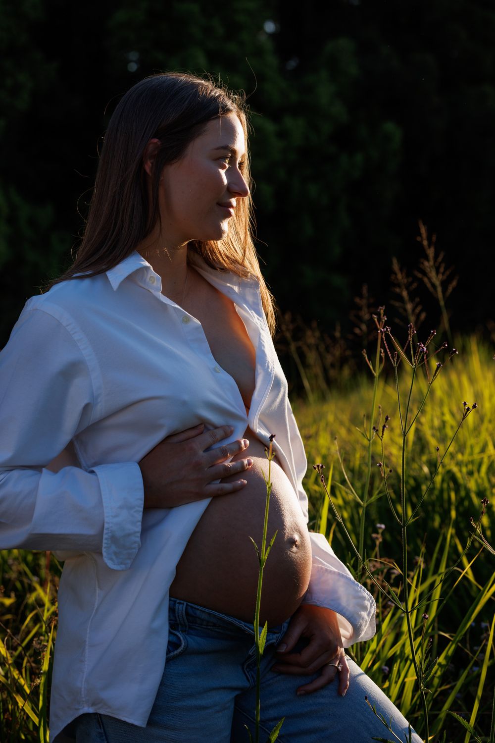 Side profile of a pregnant woman in a white button-up shirt cradling her bare belly in warm sunset light among tall grass.