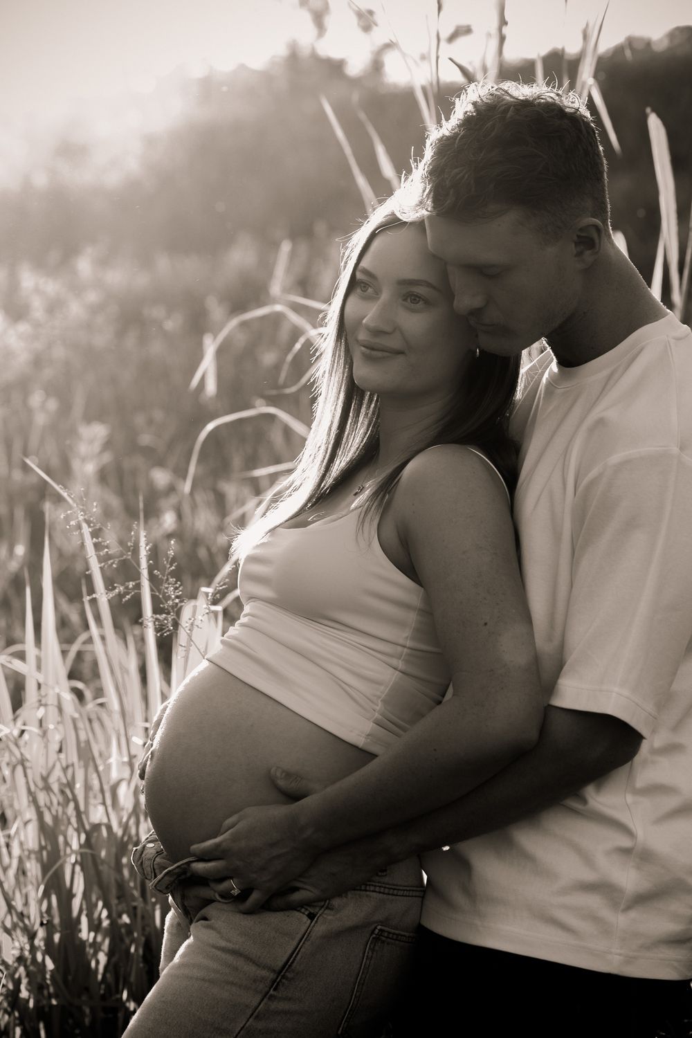 Black and white maternity photo of a man embracing his pregnant partner from behind as she smiles softly among tall grasses.