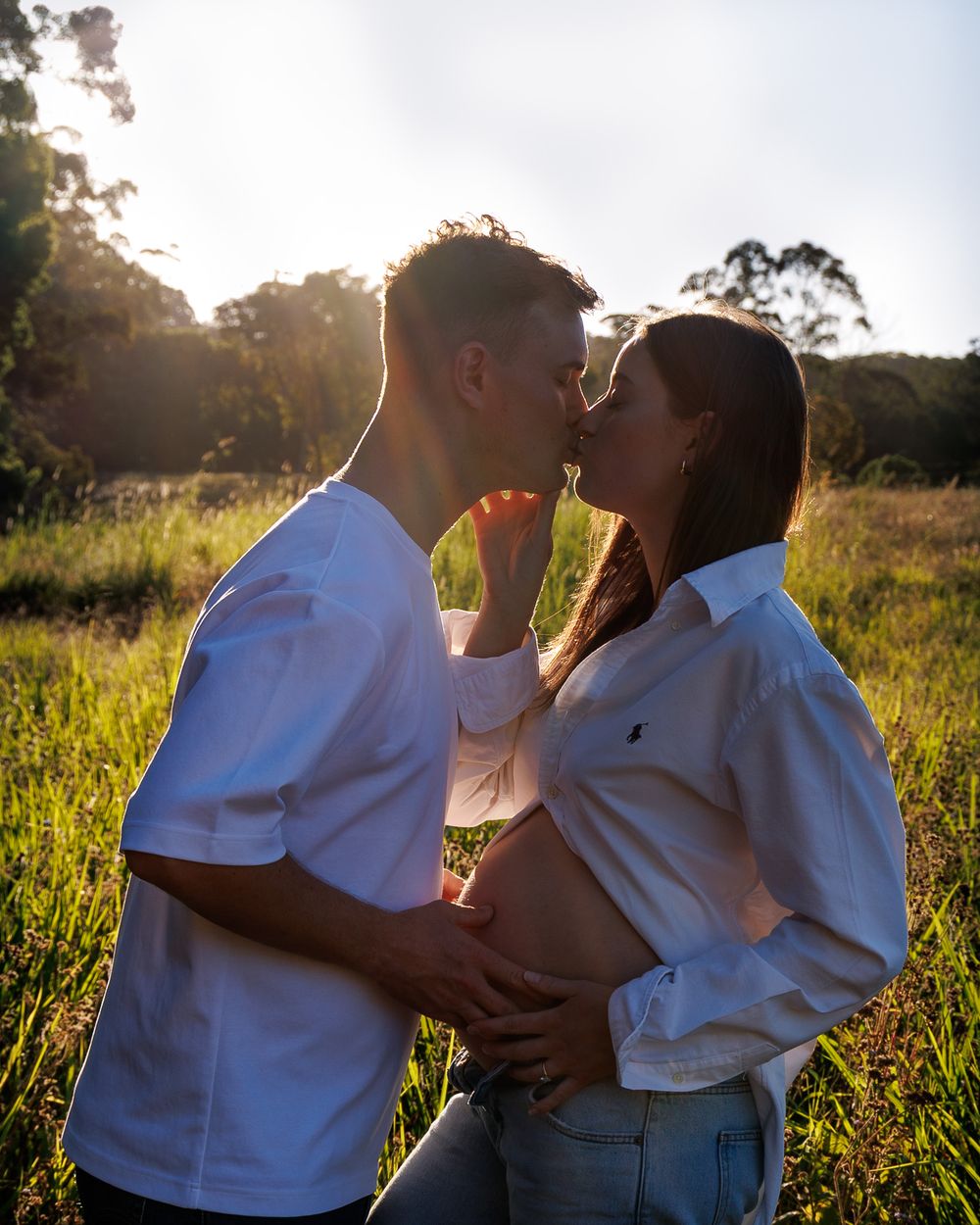 Expectant couple shares a kiss in a sunlit field, the man's hand resting on his partner's pregnant belly at golden hour.