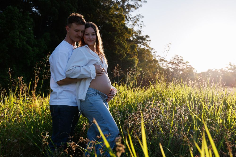 Expectant couple embracing in a sunlit grassy field at golden hour during an outdoor maternity photo session.
