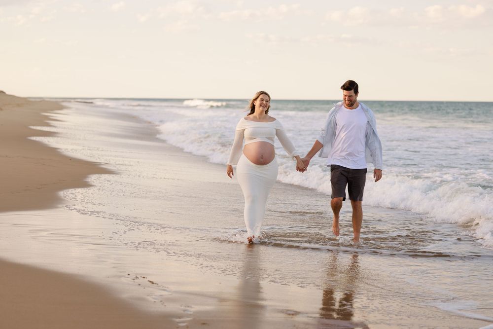 Expecting couple holding hands and walking along the shoreline at sunset in a beachside maternity photo shoot.