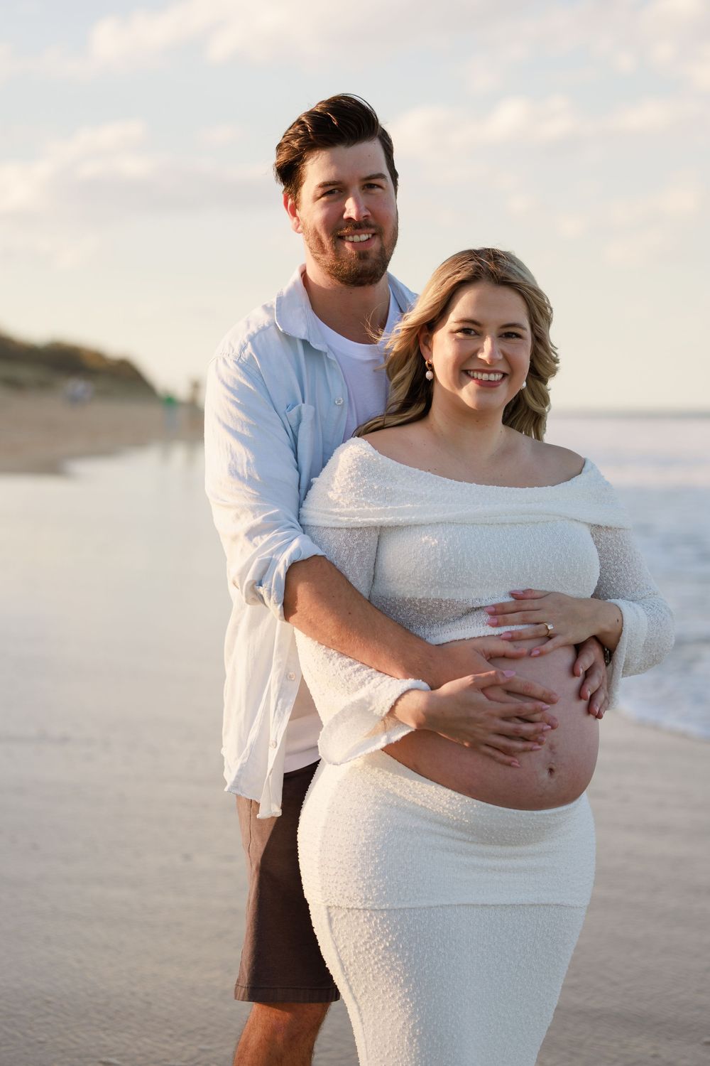 Smiling man embracing his pregnant partner from behind on the beach, both hands resting on her baby bump during maternity photos.