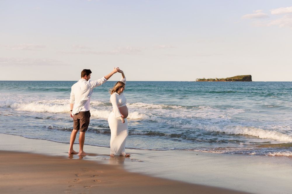 Expectant couple dancing barefoot on a sunny beach, the man twirling his pregnant partner in a white dress by the waves.