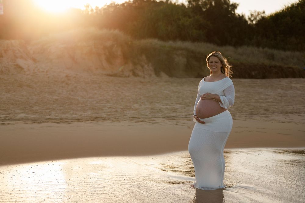 Pregnant woman in a white dress standing in shallow water at the beach as the golden sun sets behind the dunes.