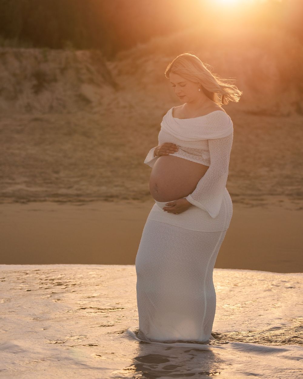 Pregnant woman in a white off-shoulder outfit stands in the surf with her hair blowing in the wind and warm sunset glow behind her.
