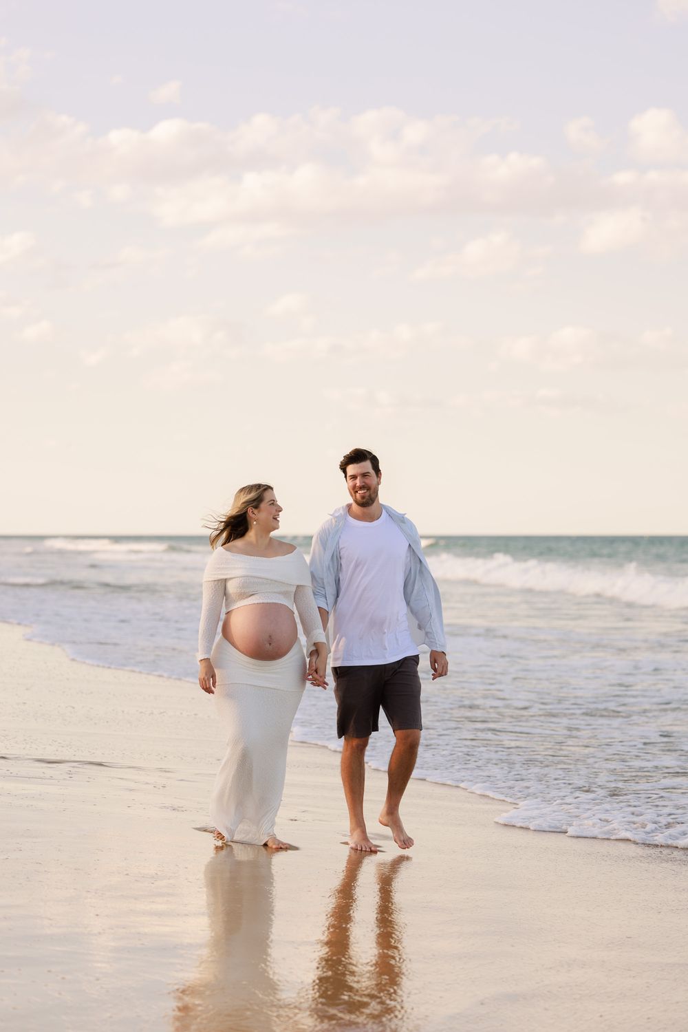 Pregnant woman in a white maternity outfit walks hand in hand with her smiling partner along the shoreline at sunset.