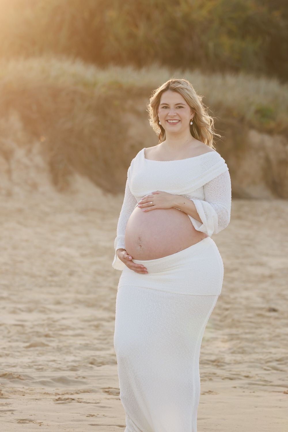 Smiling pregnant woman in an elegant white off-the-shoulder maternity dress cradles her bump on a sandy beach at golden hour.