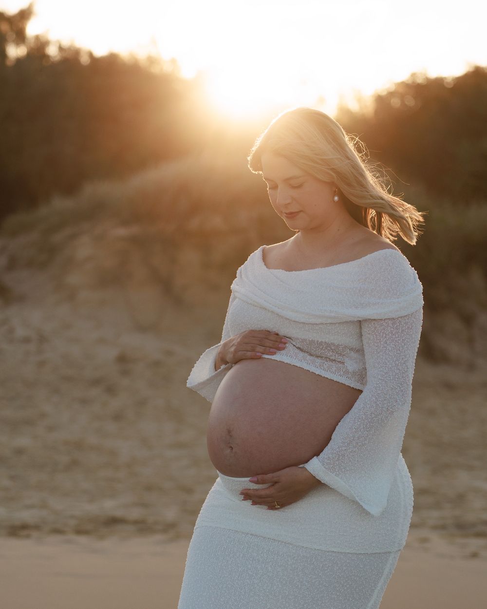 Pregnant woman in a white knit maternity outfit looks down at her belly as the setting sun glows behind her on the beach.