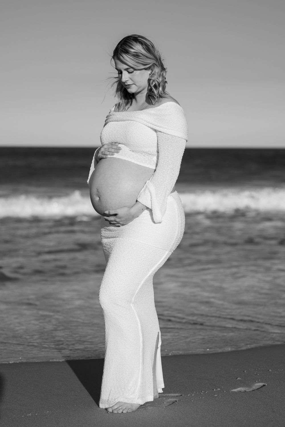 Black and white maternity portrait of a pregnant woman in a fitted white gown holding her bump by the ocean waves.