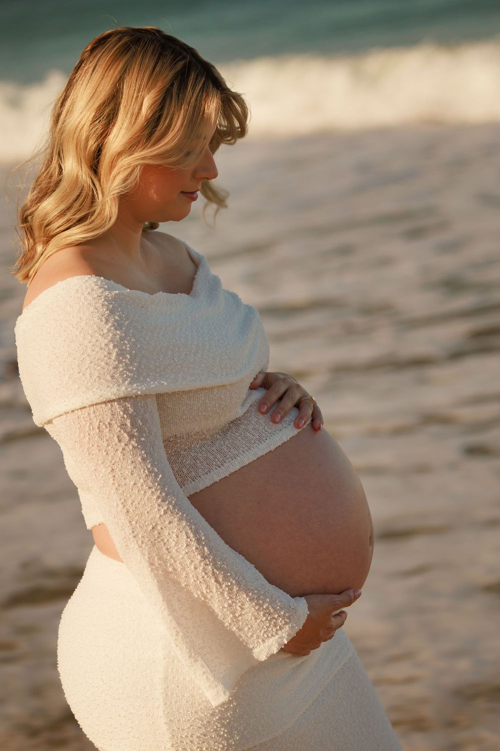 Pregnant woman in a white knit outfit cradling her baby bump while standing on a beach with ocean waves behind her.