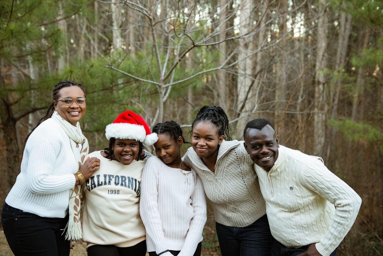 Five smiling family members in cozy cream sweaters pose close together outdoors in a wooded area, one child wearing a Santa hat.