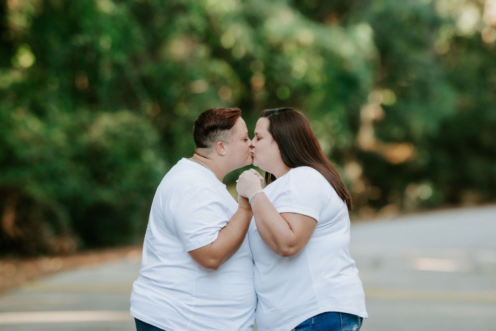 Two people in matching white t-shirts share a kiss while embracing on a tree-lined road with blurred green foliage in the background.